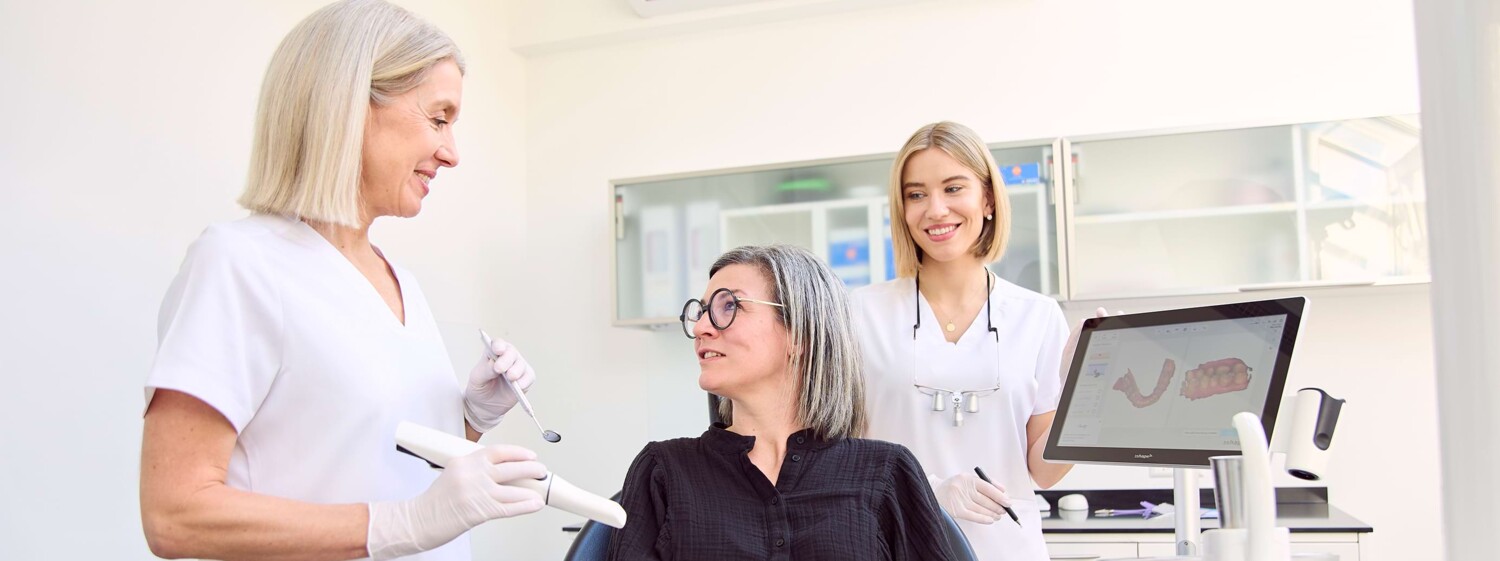 Julia Schneider und Silvia Hahn stehen neben einer Patientin im Behandlungsstuhl und erklären das Vorgehen